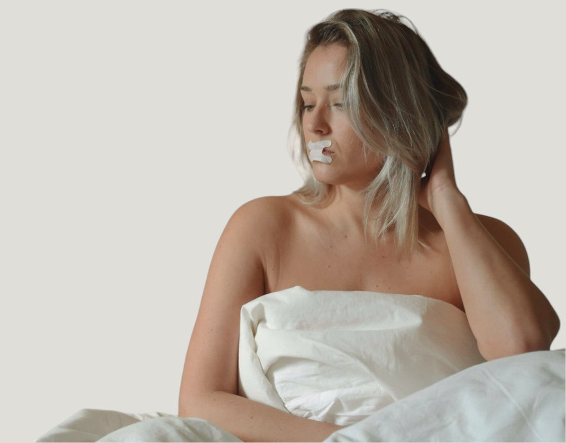 A blonde woman sits up in bed. She's wearing a Resty h shape mouthtape