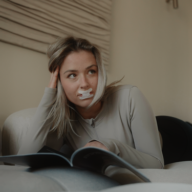 A blonde woman is reading a book. She's wearing a Resty h shape mouth tape