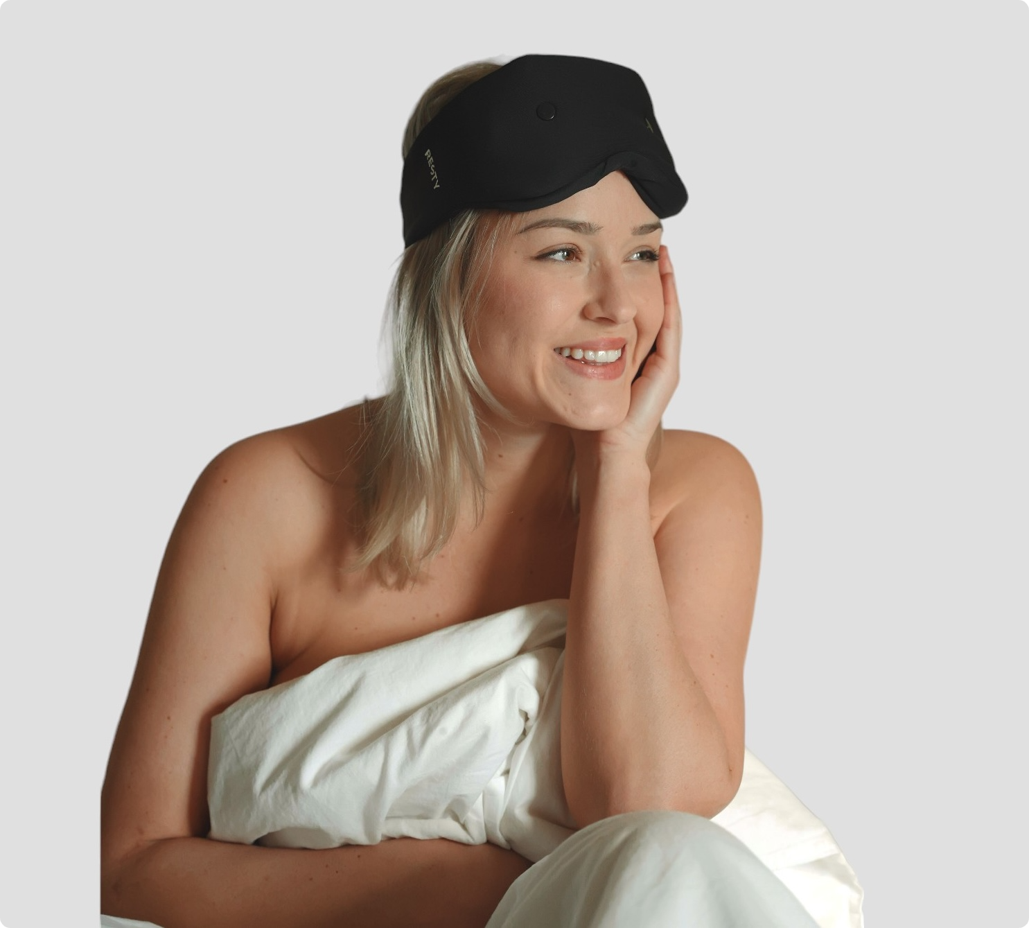 A blonde woman sits up in bed. She's wearing a Resty sleepmask